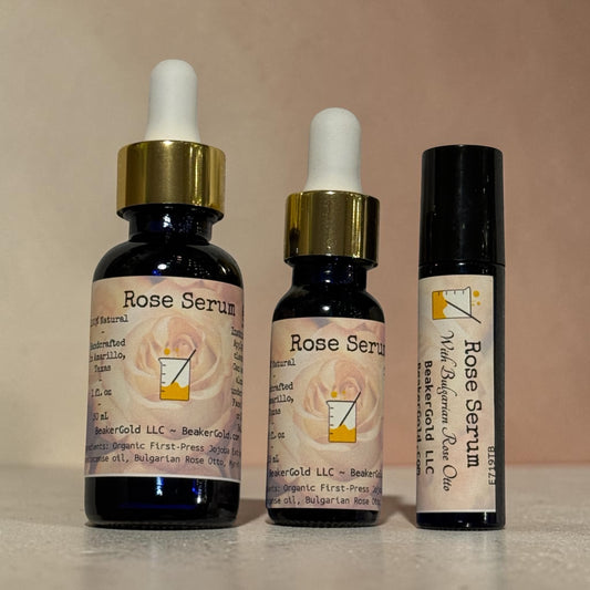 Three cosmetic containers for Rose Serum by Eterno's Rose Gold LLC are lined up against a plain beige background: a large dropper bottle, a smaller dropper bottle, and a black roller bottle with a white cap.