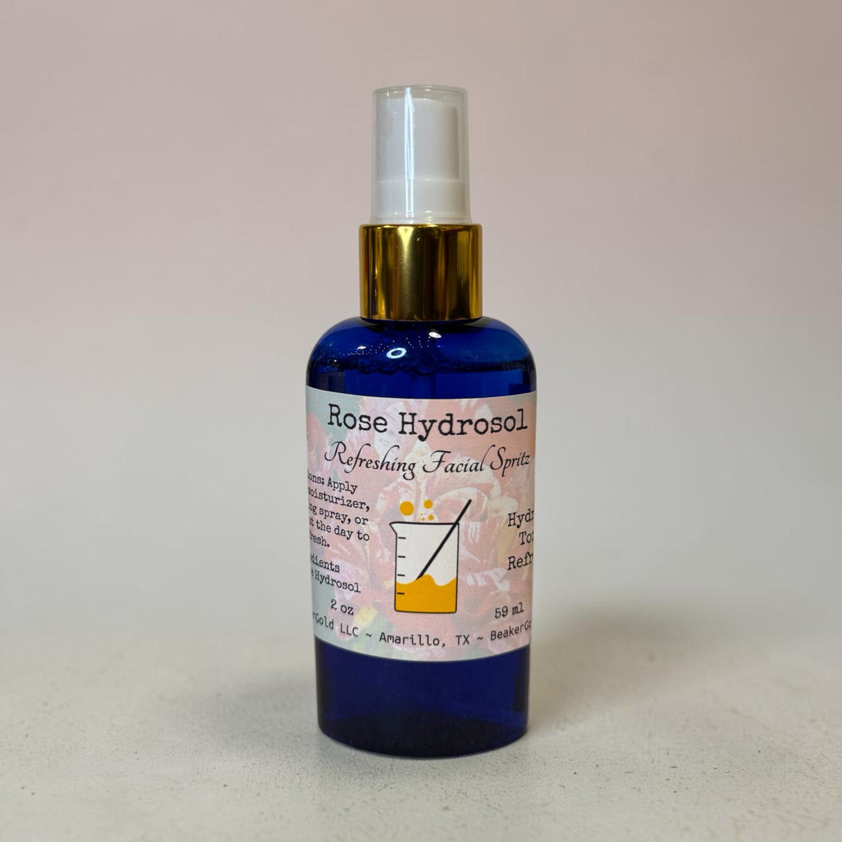 A 2 oz travel size spray bottle of Rose Hydrosol Refreshing Facial Spray, featuring a cobalt blue glass bottle, a gold collar, and a white spray nozzle, set against a plain light beige background.